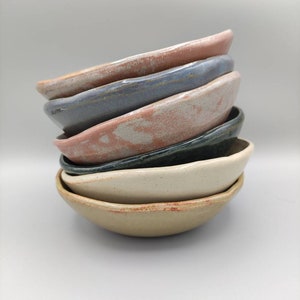 Peut inclure: Un ensemble de six bols en céramique de différentes couleurs, dont le beige, le blanc, le gris, le bleu et le rose. Les bols sont tous de formes et de tailles légèrement différentes et ont un aspect rustique et fait main.