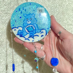 May include: A round, blue and white Care Bear suncatcher with a grumpy bear illustration. The suncatcher has silver chain and bead accents, with a price tag of "55" in the upper right corner.