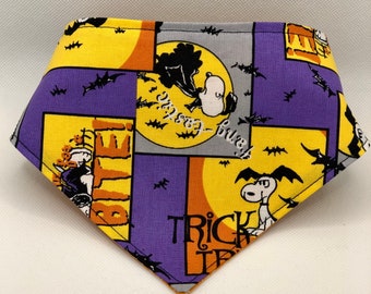 May include: A Halloween-themed dog bandana with a patchwork design. The bandana features Snoopy and Woodstock characters, bats, and the phrases "Trick or Treat" and "Fang-tastic". The color scheme includes purple, yellow, orange, and gray.