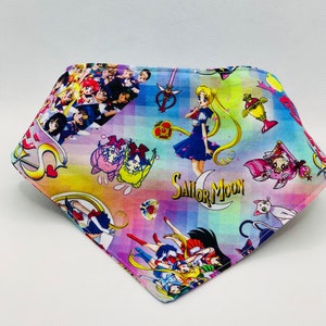May include: A colourful bandana featuring a Sailor Moon theme. The bandana has a rainbow gradient background with a checkered pattern. The bandana is covered with images of Sailor Moon characters, including Sailor Moon, Sailor Mercury, Sailor Mars, Sailor Jupiter, and Sailor Venus. The bandana also features the Sailor Moon logo and other Sailor Moon-related imagery.