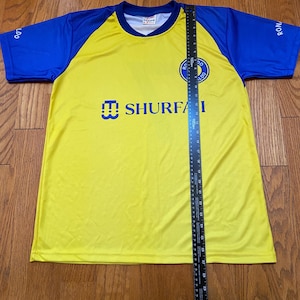 Peut inclure: Maillot de football jaune et bleu avec le texte "SHURFA" et un logo d'&eacute;quipe sur la poitrine. Les manches sont bleues, et le maillot est mesur&eacute; avec une r&egrave;gle.