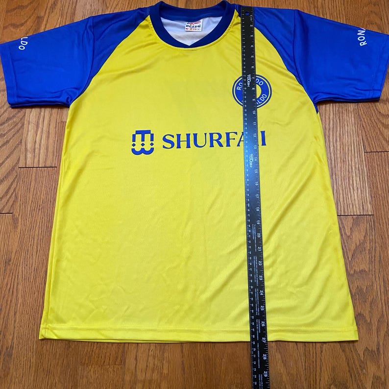 Peut inclure: Maillot de football jaune et bleu avec le texte "SHURFA" et un logo d'&eacute;quipe sur la poitrine. Les manches sont bleues, et le maillot est mesur&eacute; avec une r&egrave;gle.