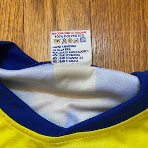 Peut inclure: Gros plan d'un maillot jaune et bleu avec une &eacute;tiquette blanche. L'&eacute;tiquette indique "NO CONFORME AL ORIGINAL 100% POLYESTER" et comprend des instructions de lavage en espagnol. Le col du maillot est bleu.