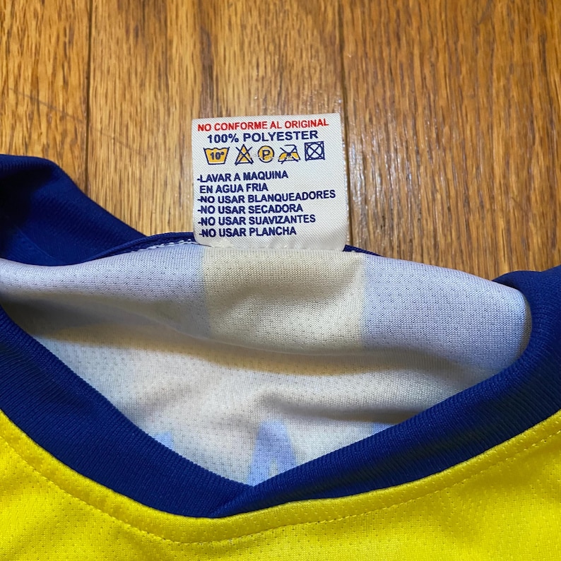 Peut inclure: Gros plan d'un maillot jaune et bleu avec une &eacute;tiquette blanche. L'&eacute;tiquette indique "NO CONFORME AL ORIGINAL 100% POLYESTER" et comprend des instructions de lavage en espagnol. Le col du maillot est bleu.