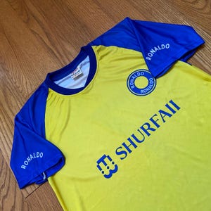 Peut inclure: Maillot de football jaune et bleu avec le nom "RONALDO" imprim&eacute; sur les manches et un logo circulaire sur la poitrine. Le maillot porte &eacute;galement le texte "SHURFAII" en bleu. Col rond et manches courtes.
