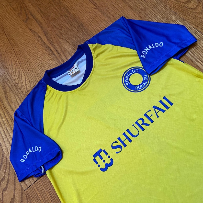Peut inclure: Maillot de football jaune et bleu avec le nom "RONALDO" imprim&eacute; sur les manches et un logo circulaire sur la poitrine. Le maillot porte &eacute;galement le texte "SHURFAII" en bleu. Col rond et manches courtes.
