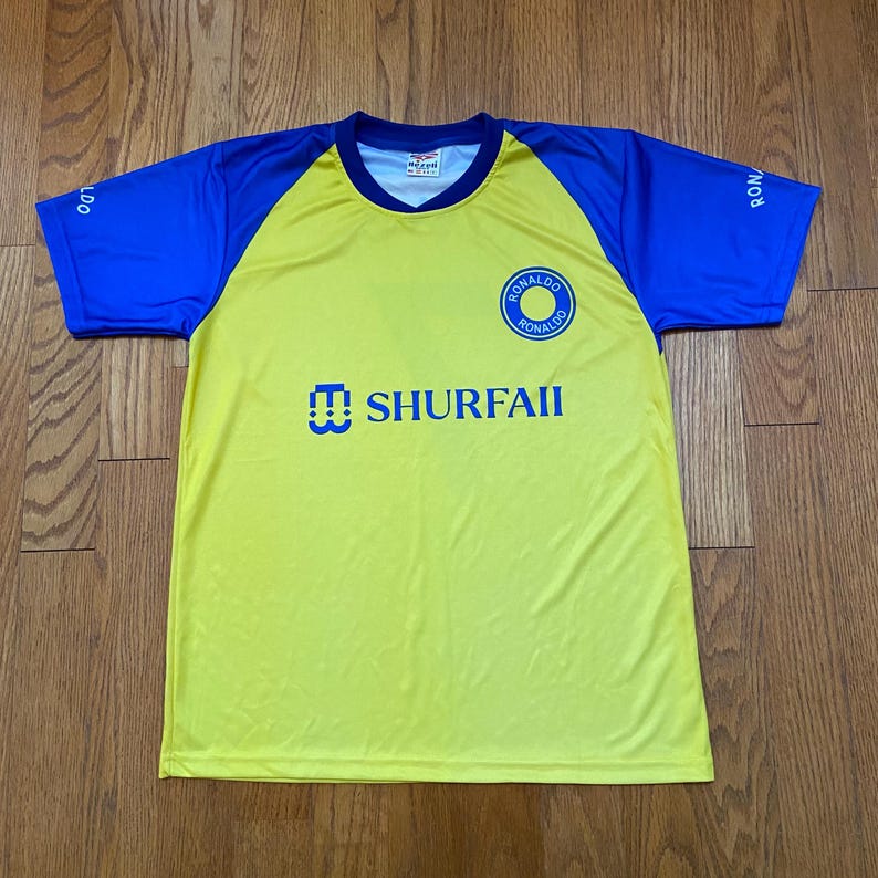 Peut inclure: Maillot de football jaune et bleu avec le texte "SHURFAII" et un logo circulaire indiquant "RONALDO". Les manches et les &eacute;paules sont bleues, et le corps est jaune. Le maillot a un col en V.