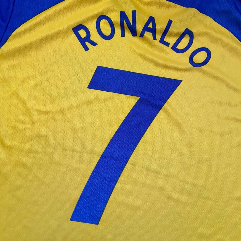 Peut inclure: Gros plan sur un maillot de football jaune avec des d&eacute;tails bleus. Le nom "RONALDO" est imprim&eacute; en bleu au-dessus du num&eacute;ro "7". Le maillot a une texture.