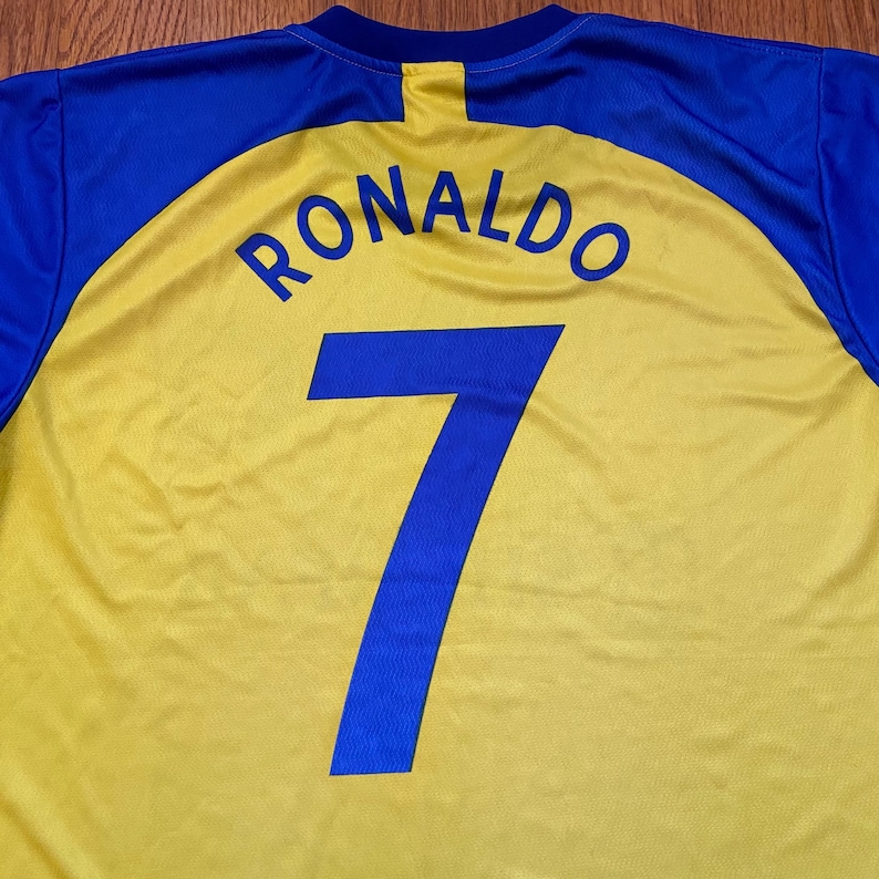 Peut inclure: Un maillot de football jaune et bleu avec le nom "RONALDO" et le num&eacute;ro "7" en bleu. Le maillot a un corps jaune avec des &eacute;paules et des manches bleues.