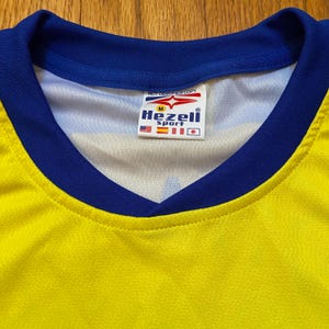 Peut inclure: Maillot de sport jaune et bleu avec un col en V. Le col et les bords des manches sont bleus. Une &eacute;tiquette &agrave; l'int&eacute;rieur du col indique "Hezeli Sport" avec les drapeaux des &Eacute;tats-Unis, d'Espagne et du Japon. Le texte "NOT CERTIFIED COPY" est au-dessus de l'&eacute;tiquette.