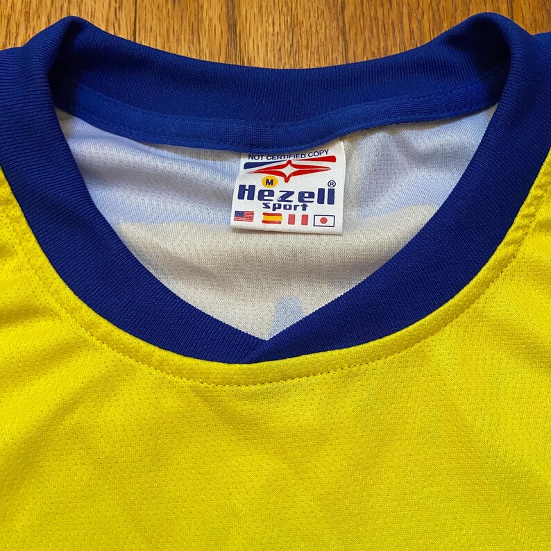 Peut inclure: Maillot de sport jaune et bleu avec un col en V. Le col et les bords des manches sont bleus. Une &eacute;tiquette &agrave; l'int&eacute;rieur du col indique "Hezeli Sport" avec les drapeaux des &Eacute;tats-Unis, d'Espagne et du Japon. Le texte "NOT CERTIFIED COPY" est au-dessus de l'&eacute;tiquette.
