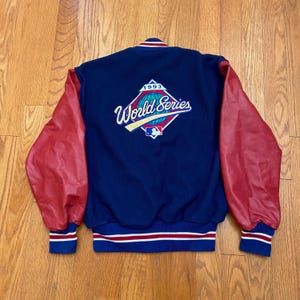 May include: A vintage baseball jacket with a navy blue body and red sleeves. The back features a "1993 World Series" emblem. The jacket has blue, red, and white striped trim at the waist and cuffs. A classic piece of sports apparel.