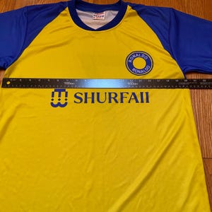Peut inclure: Maillot de football jaune et bleu avec le texte "SHURFAII" et un embl&egrave;me circulaire avec "RONALDO" en bleu. Le maillot a des manches bleues et un col en V.