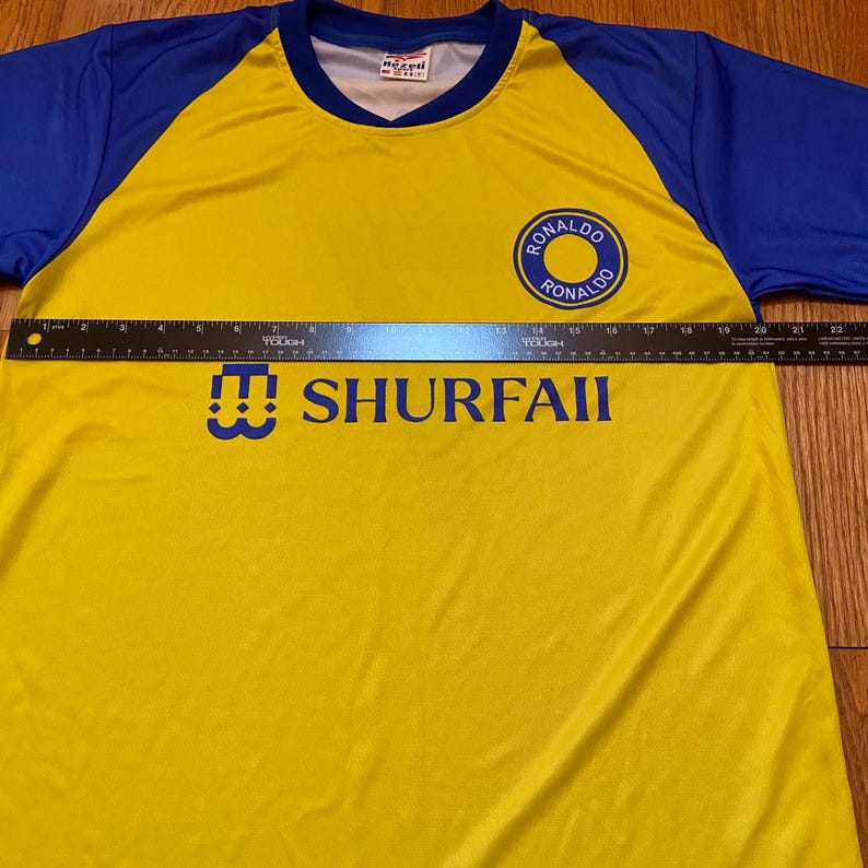 Peut inclure: Maillot de football jaune et bleu avec le texte "SHURFAII" et un embl&egrave;me circulaire avec "RONALDO" en bleu. Le maillot a des manches bleues et un col en V.