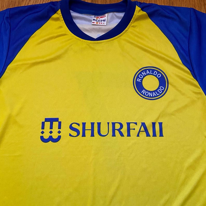 Peut inclure: Maillot de football jaune et bleu avec le texte "SHURFAII" en bleu. Le maillot pr&eacute;sente un &eacute;cusson circulaire avec "RONALDO" en bleu. Les manches et le col sont bleus.