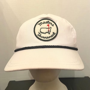 Può includere: Un cappellino da baseball bianco con una toppa circolare sul davanti. La toppa mostra il contorno nero degli Stati Uniti con una bandierina da golf rossa e la scritta Masters Tournament in corsivo nero. Un cordoncino nero intrecciato attraversa la visiera.