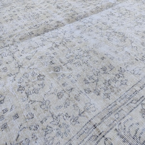 May include: Close-up of a vintage beige rug with a faded floral pattern in shades of gray and brown. The rug has a distressed look, with visible wear and tear, adding to its antique charm. The rug's design features a central field with floral motifs and a border.