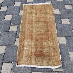 May include: A rectangular, distressed-look rug with a faded, warm color palette of brown and orange. The rug features a central medallion design and a fringed edge. The rug is placed on a gray and white stone surface.