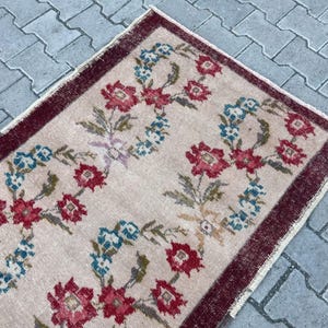 Pu&ograve; includere: Un tappeto rettangolare con sfondo beige e motivo floreale. Il design presenta fiori rossi e blu disposti simmetricamente, con un bordo rosso scuro. Il tappeto ha un aspetto vintage e usurato.