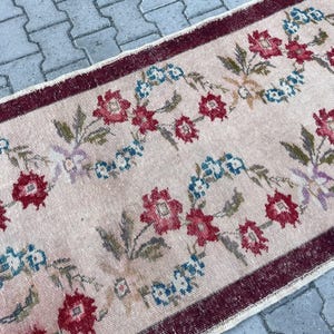 Pu&ograve; includere: Un tappeto rettangolare con sfondo rosa chiaro e motivo floreale. Il design presenta fiori rossi e blu con foglie verdi, incorniciati da un bordo rosso scuro. Il tappeto ha un aspetto vintage e usurato.