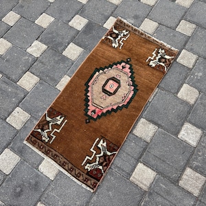 May include: A rectangular rug featuring a brown field with a central geometric medallion in beige, pink, and dark green. The rug includes four corner motifs with white and dark brown patterns and short white fringe on both ends.