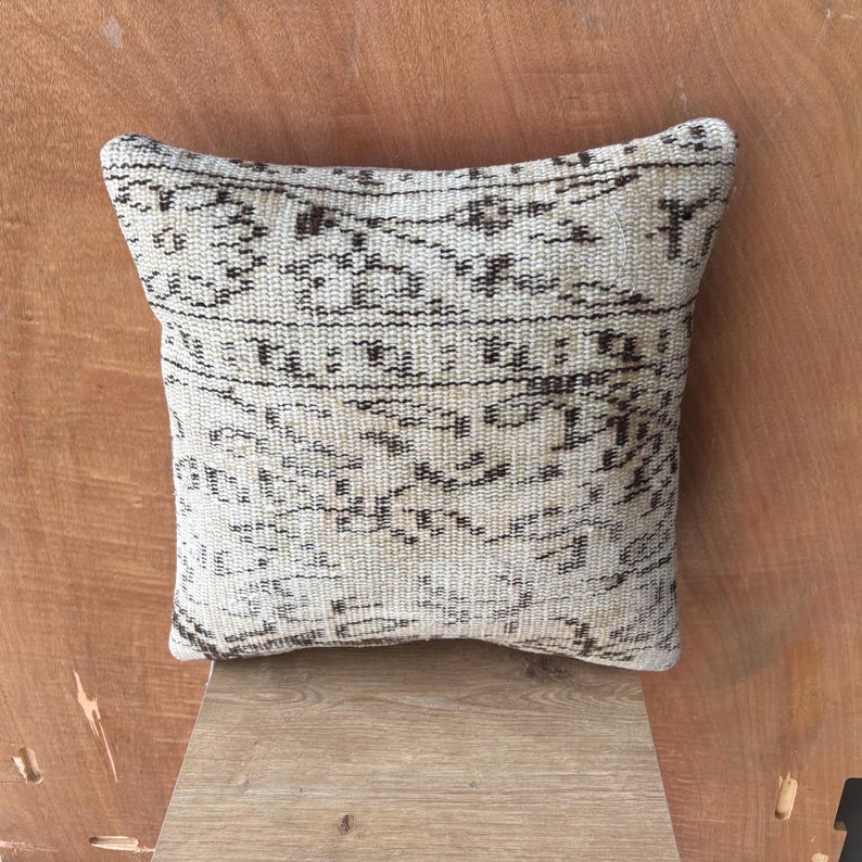 May include: A square, cream-coloured cushion featuring a dark brown, detailed pattern. The cushion has a gently curved shape and is made from woven fabric. The design appears to be a traditional textile pattern, possibly vintage.