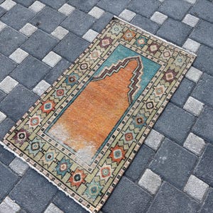 Puede incluir: Una alfombra rectangular que presenta un diseño central de mihrab naranja sobre un fondo verde azulado, rodeada por un borde ancho con patrones geométricos en tonos naranja, verde azulado y marrón, con flecos blancos cortos en los bordes.