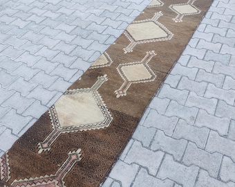 Peut inclure: Un long tapis de couloir rectangulaire avec un motif géométrique. Le tapis présente des losanges beiges et bruns répétés, avec une bordure marron foncé et des accents roses. Le tapis est fait d'une matière tissée.