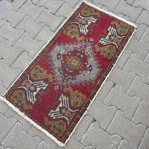 May include: A rectangular woven rug featuring a deep red background with a central grey diamond medallion. The design includes geometric patterns in olive green, orange, and white, finished with short white fringe along the two shorter edges.