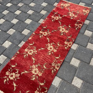 Puede incluir: Una alfombra rectangular roja con un estampado floral en beige y marrón. La alfombra tiene un aspecto desgastado, con desgaste visible. El diseño floral se repite a lo largo de la alfombra.