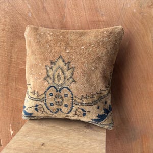 May include: A square, tan cushion with a detailed, vintage-style design in shades of blue and beige. The cushion features a central floral motif and ornate border patterns, suggesting a rug-inspired aesthetic. The cushion is placed on a wooden surface.