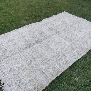 May include: A rectangular, off-white rug with a faded floral pattern and fringed edges. The rug is laid out on a bed of green grass, showcasing its neutral color palette and vintage design. The rug's dimensions are not visible.