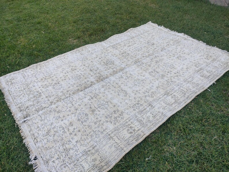 May include: A rectangular, off-white rug with a faded floral pattern and fringed edges. The rug is laid out on a bed of green grass, showcasing its neutral color palette and vintage design. The rug's dimensions are not visible.