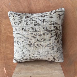 May include: Square beige cushion with a dark brown, intricate pattern. The cushion has a woven texture and a vintage aesthetic, suitable for home decor. The cushion is placed on a wooden surface.