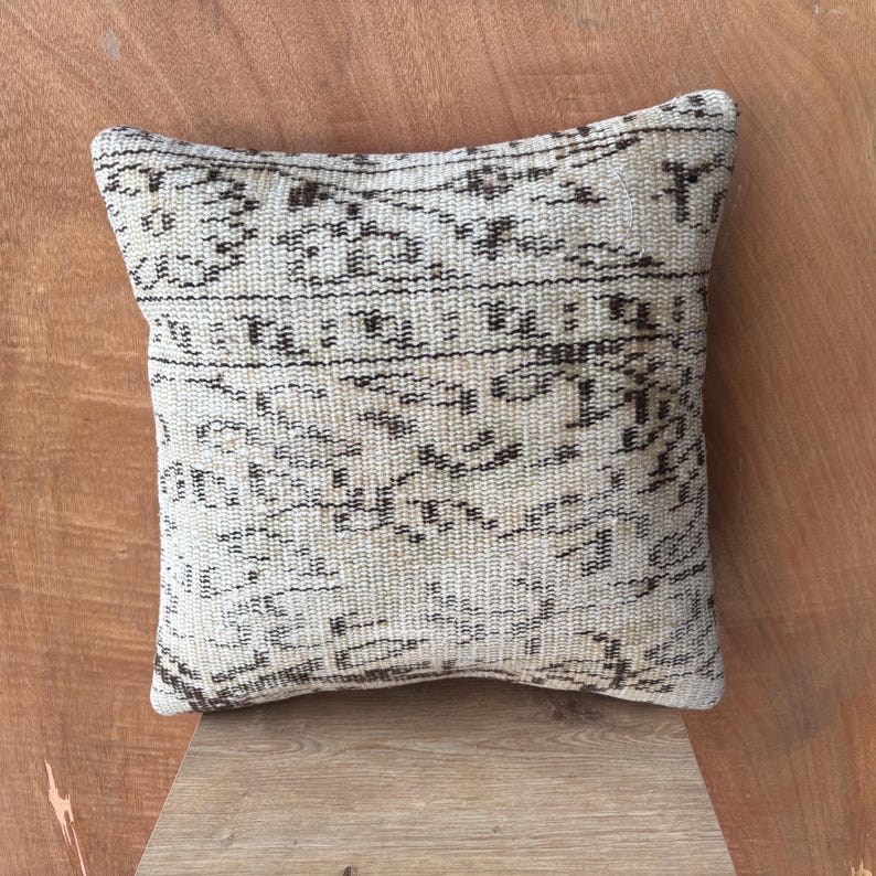 May include: Square beige cushion with a dark brown, intricate pattern. The cushion has a woven texture and a vintage aesthetic, suitable for home decor. The cushion is placed on a wooden surface.