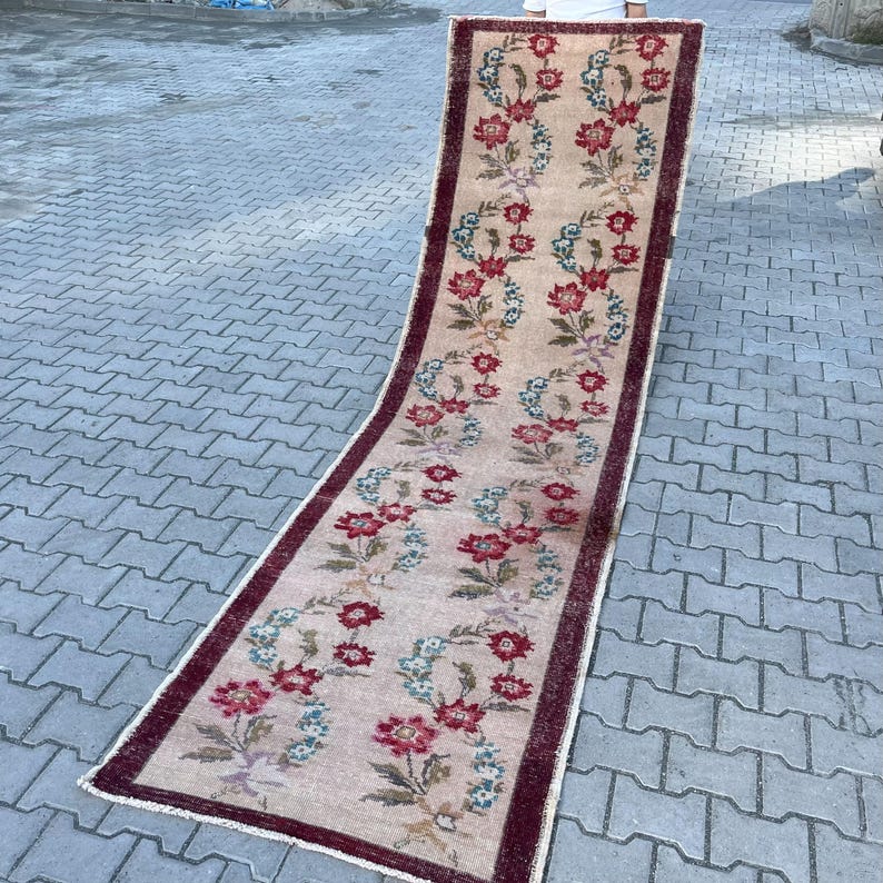 Pu&ograve; includere: Un lungo tappeto rettangolare con un motivo floreale. Il tappeto ha uno sfondo beige con un motivo ripetuto di fiori rossi e blu. Il bordo &egrave; di colore bordeaux scuro. Il tappeto &egrave; probabilmente realizzato in materiale tessuto.