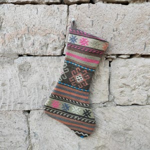 May include: A colorful, patterned Christmas stocking with a loop for hanging. The stocking features a geometric design in shades of brown, pink, blue, green, and orange. The stocking is made of a woven fabric and hangs against a stone wall.