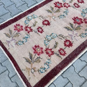 Pu&ograve; includere: Un tappeto rettangolare con un motivo floreale. Il tappeto ha uno sfondo beige chiaro con un bordo rosso scuro. Il motivo floreale comprende fiori rossi e blu con foglie verdi.