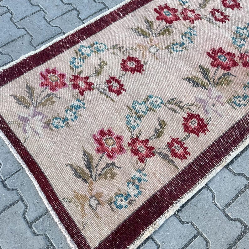 Pu&ograve; includere: Un tappeto rettangolare con un motivo floreale. Il tappeto ha uno sfondo beige chiaro con un bordo rosso scuro. Il motivo floreale comprende fiori rossi e blu con foglie verdi.
