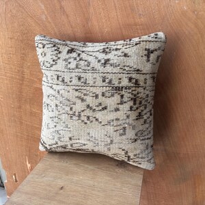 May include: Square beige cushion with a dark brown, detailed pattern. The cushion has a woven texture and appears to be made of natural fibres. The design is a traditional, possibly vintage, style. The cushion is placed on a wooden surface.