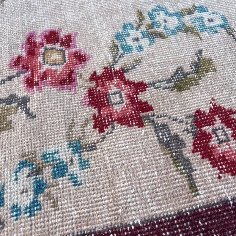 Pu&ograve; includere: Primo piano di un tappeto beige con un motivo floreale. Il tappeto presenta fiori rossi, rosa e blu con foglie verdi. Il bordo &egrave; di colore bordeaux scuro. Il tappeto ha un aspetto vintage e fatto a mano.