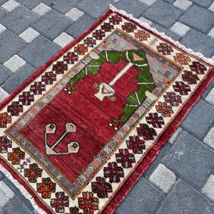 May include: A rectangular area rug featuring a deep red central field with a green arch motif and a white floral-like emblem. The border displays a repeating pattern of geometric floral shapes in red, green, and orange on a cream background.