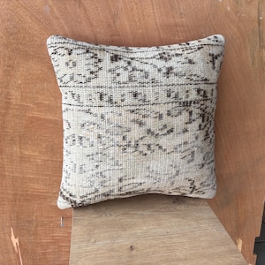 May include: Square beige cushion with a woven, distressed pattern in dark brown. The cushion has a vintage, textured appearance, suggesting a handmade or antique origin. The design features geometric and abstract shapes.