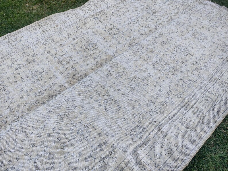 May include: A rectangular, vintage-style rug with a faded, floral pattern in shades of cream and gray. The rug has a distressed look, with visible wear and tear, and is laid out on a grassy surface.