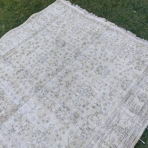 May include: A rectangular, off-white rug with a faded floral pattern and a distressed look. The rug has a border with a repeating design and fringe along one edge. The rug is lying on green grass.