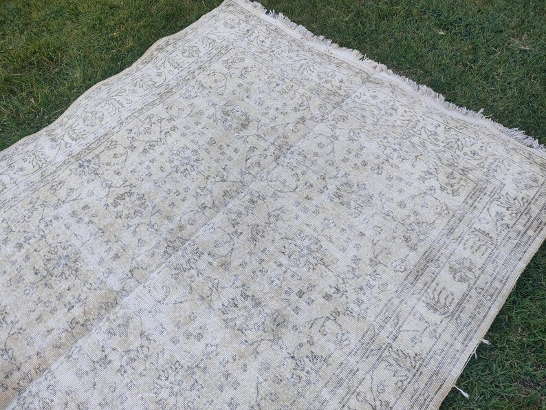 May include: A rectangular, off-white rug with a faded floral pattern and a distressed look. The rug has a border with a repeating design and fringe along one edge. The rug is lying on green grass.