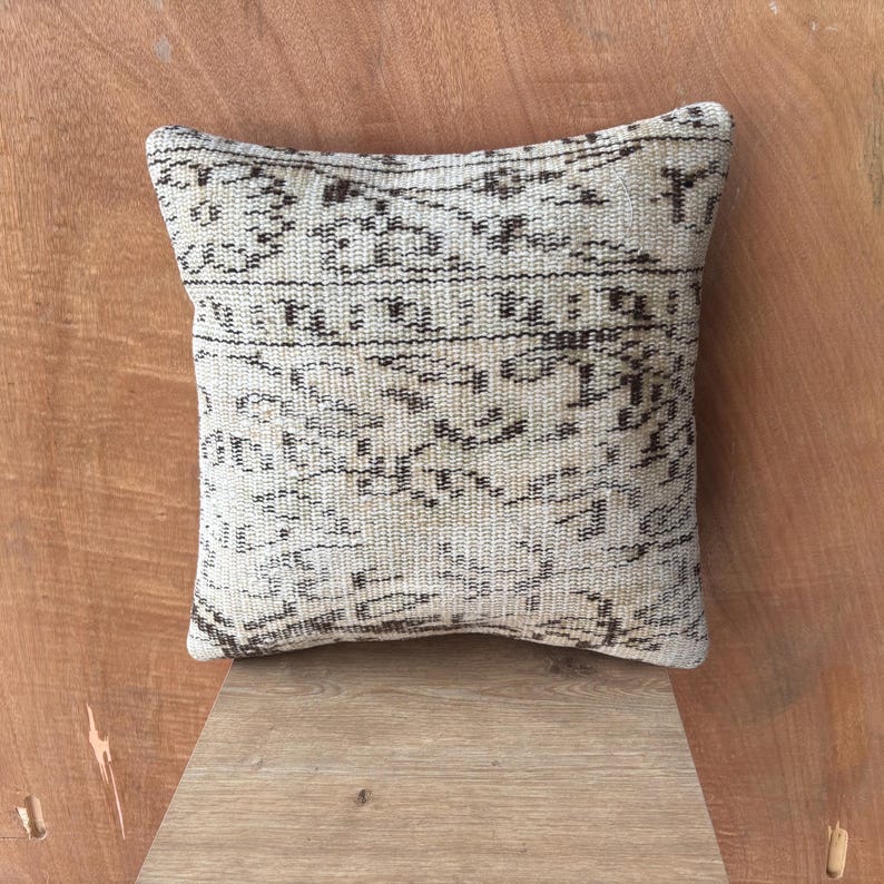 May include: A square cushion in off-white with a faded pattern in brown and black. The cushion has a textured look, possibly woven. The design suggests a traditional or vintage style, with a worn appearance.