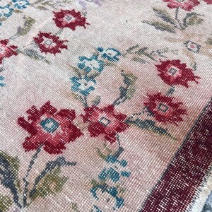 Pu&ograve; includere: Primo piano di un tappeto vintage con motivo floreale. Il tappeto presenta uno sfondo rosa chiaro con disegni floreali rossi, verde acqua e verdi. Il bordo &egrave; rosso scuro. Il tappeto ha un aspetto invecchiato e antico.