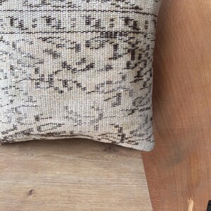 May include: Close-up of a beige and brown patterned cushion. The cushion has a woven texture with a repeating geometric design in shades of brown against a beige background. The cushion is resting on a light brown wooden surface.