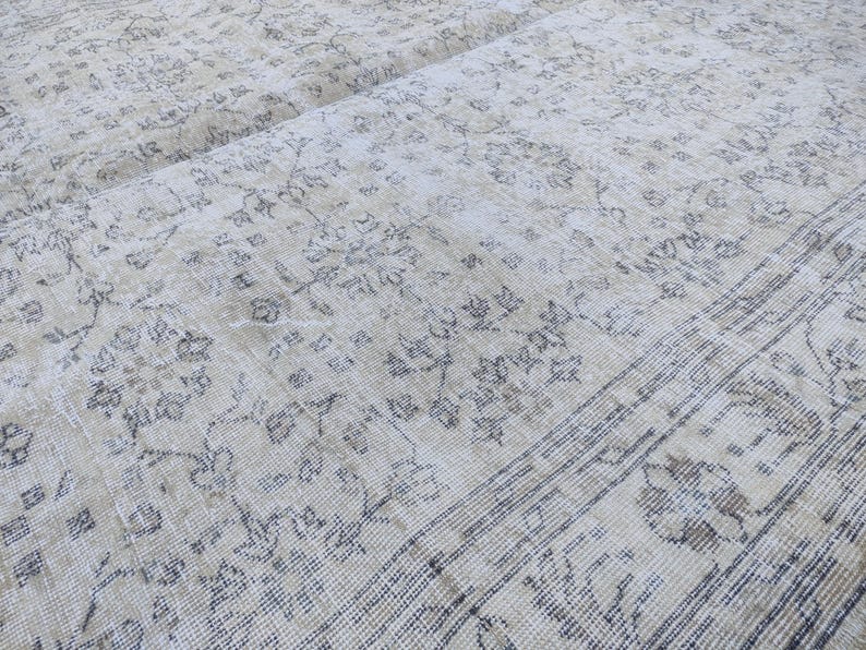 May include: Close-up of a vintage beige rug with a faded floral pattern in shades of gray and brown. The rug has a distressed look, with visible wear and tear, adding to its antique charm. The rug's design features a central field with floral motifs and a border.